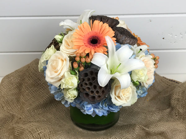 A compact vase of hydrangea, lilies, roses, Gerber daisies, and textured pods. In soft tones of whites, blues and peaches.
