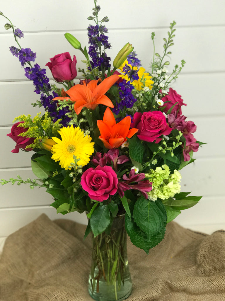 tall vase with wildflowers that include lilies, Larkspur, Gerber daisies, roses, and green hydrangea
