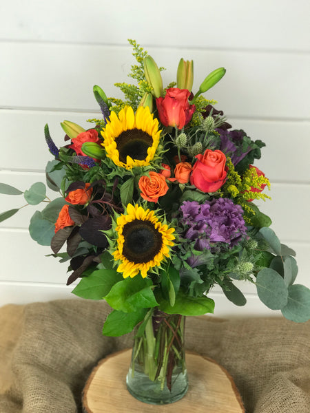 Fall collection of purples, oranges, yellows and reds. Includes Lilies, sunflowers, roses, spray roses, purple kale in a tall vase