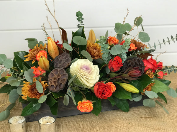 A natural wooden box is used beautifully as the vessel for this centerpiece. Filled with hues of the Fall and textures found throughout nature, it is a perfect adornment to your Thanksgving Day table.  The long and low centerpiece sits in a 18" wooden box which contains florals such as roses, spray roses, kale, hypericum, mums, lilies, leucadendron, magnolia, and eucalyptus. A stunner, to say the least! by gig Morris florist in belmar, New Jersey